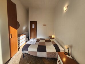 A small, neatly arranged bedroom with a checkered bedspread, a wooden wardrobe, a bedside table with a lamp, and a wall-mounted light fixture. - TATA HOMES B&B (Bologna)