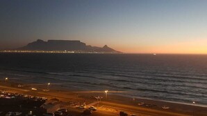 A coastal road with a view of a mountain and a city skyline in the distance. - Ocean Pearl -  iconic views of Table Mountain & Robben Island. (Cape Town)