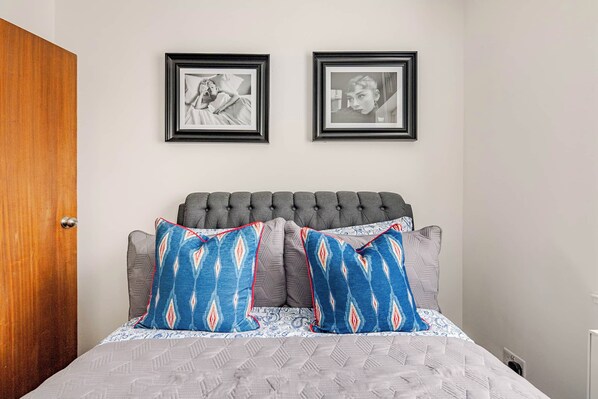 A bedroom with a tufted headboard, two framed pictures on the wall, and patterned pillows.