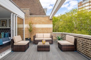 A balcony with wicker furniture, a potted plant, and a view of a brick wall and greenery.