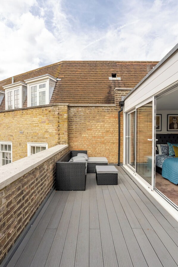A balcony with a wooden floor, a brick wall, and a sliding glass door leading to a bedroom.