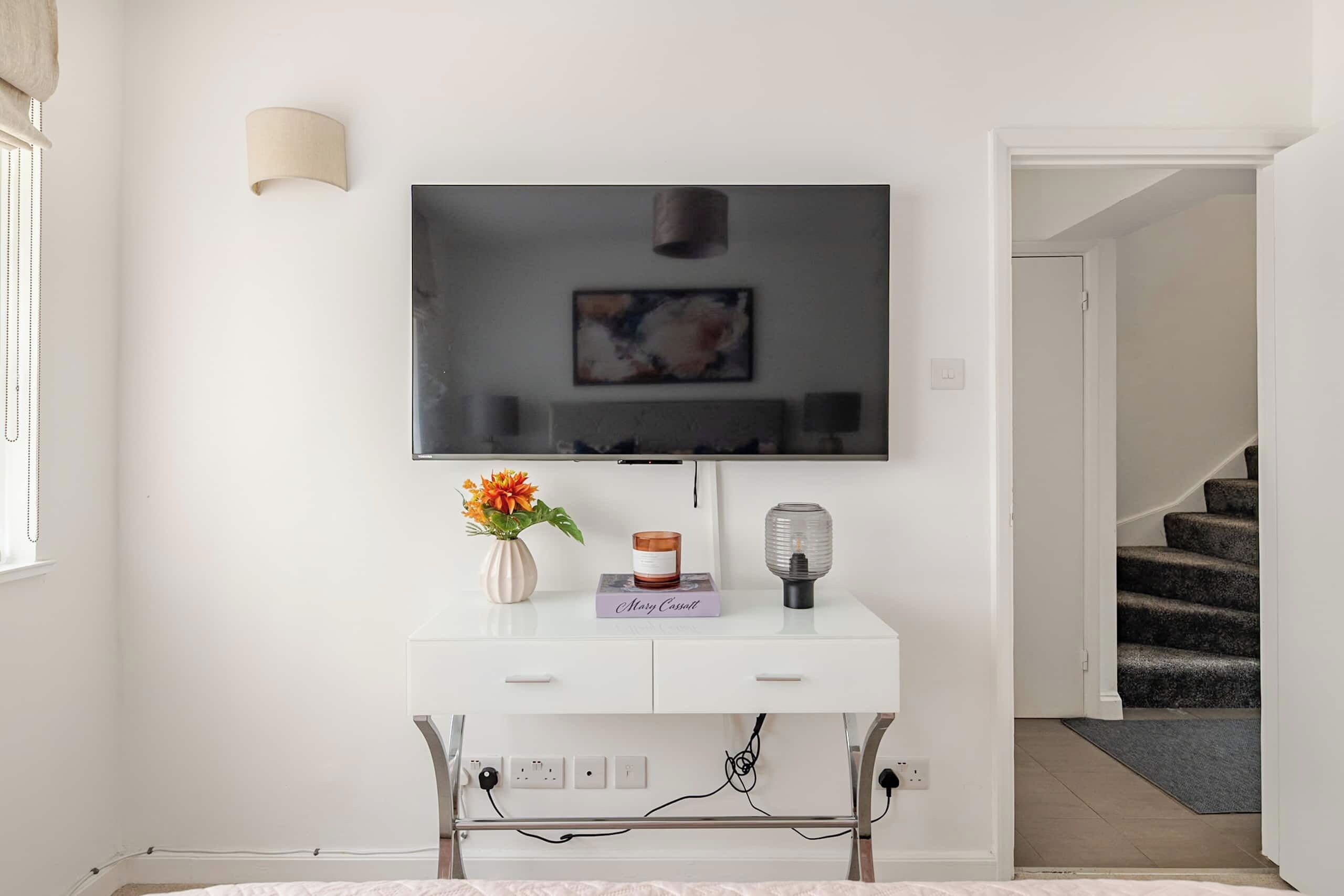 Un salon moderne avec un téléviseur à écran plat fixé sur une console blanche, un vase avec des fleurs, un livre et une lampe.