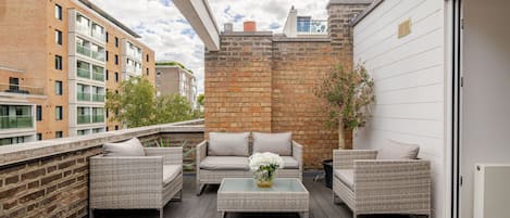 Un balcon agrémenté de meubles en osier, d’une table en verre et d’un mur en briques.
