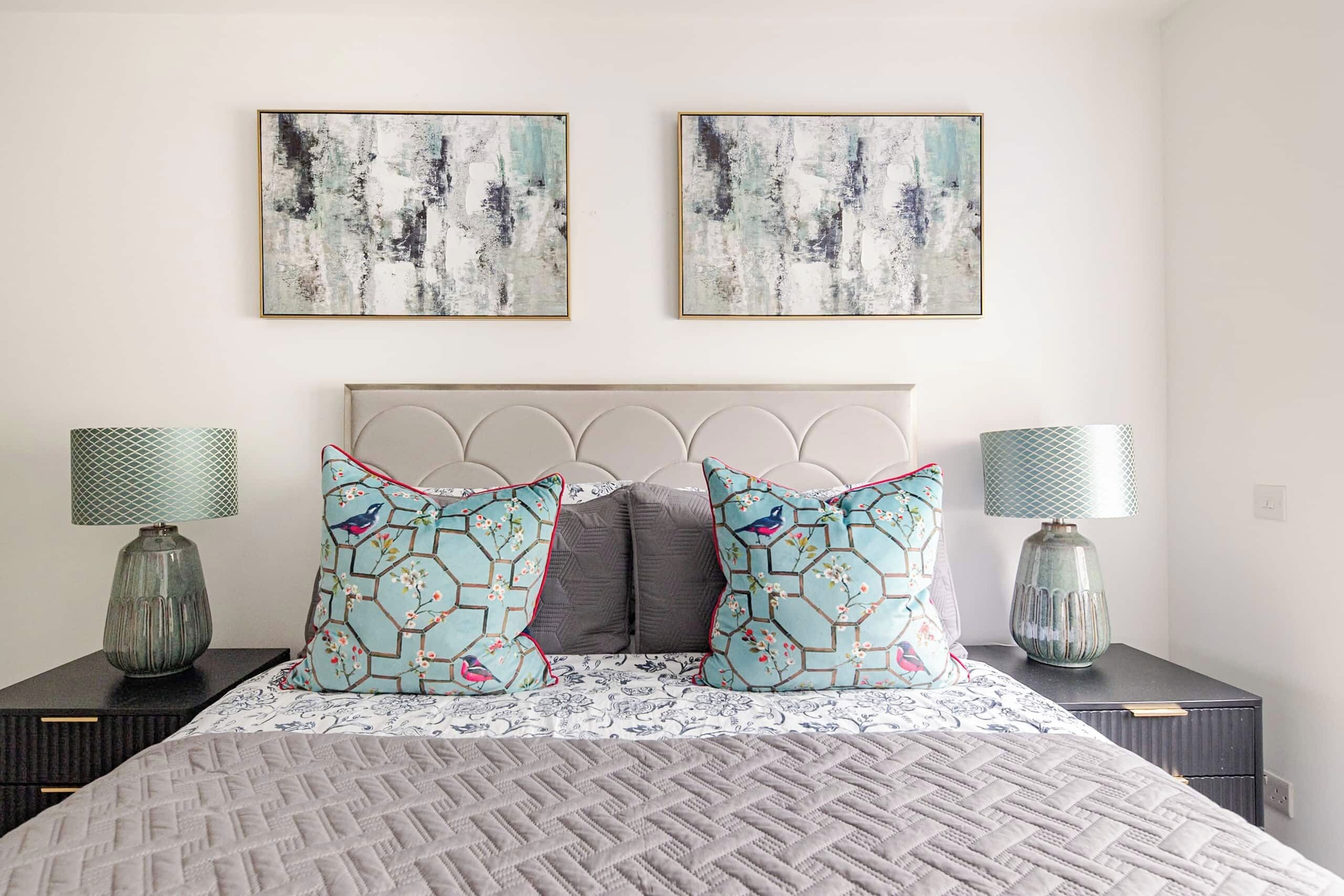 A bedroom with a bed, two nightstands, two lamps, and two abstract paintings on the wall.