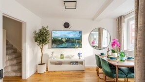 Ein modernes Wohnzimmer mit Holzboden, einer weißen Unterhaltungsecke, einem großen Flachbildfernseher, einem runden Spiegel und einem Essbereich mit grünen Stühlen.