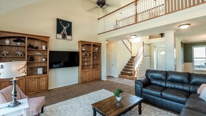 A spacious living room with a high ceiling, a wooden entertainment center, a black leather sofa, and a staircase leading to an upper level. - Charming farmhouse in Countryside with Pool & great views for 16 guests  (Ponder)