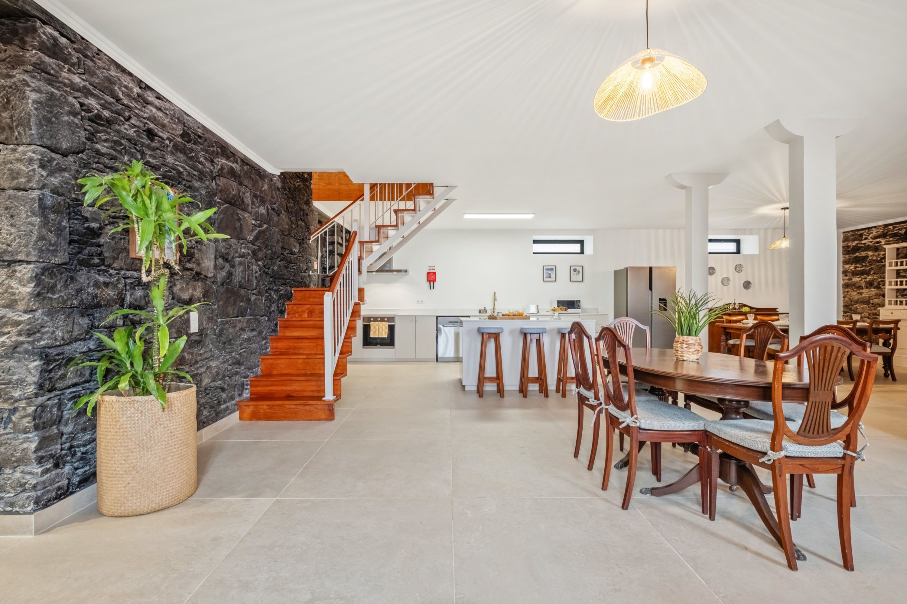 A spacious living area with a stone wall, a wooden staircase, a dining table with chairs, and a kitchen in the background.