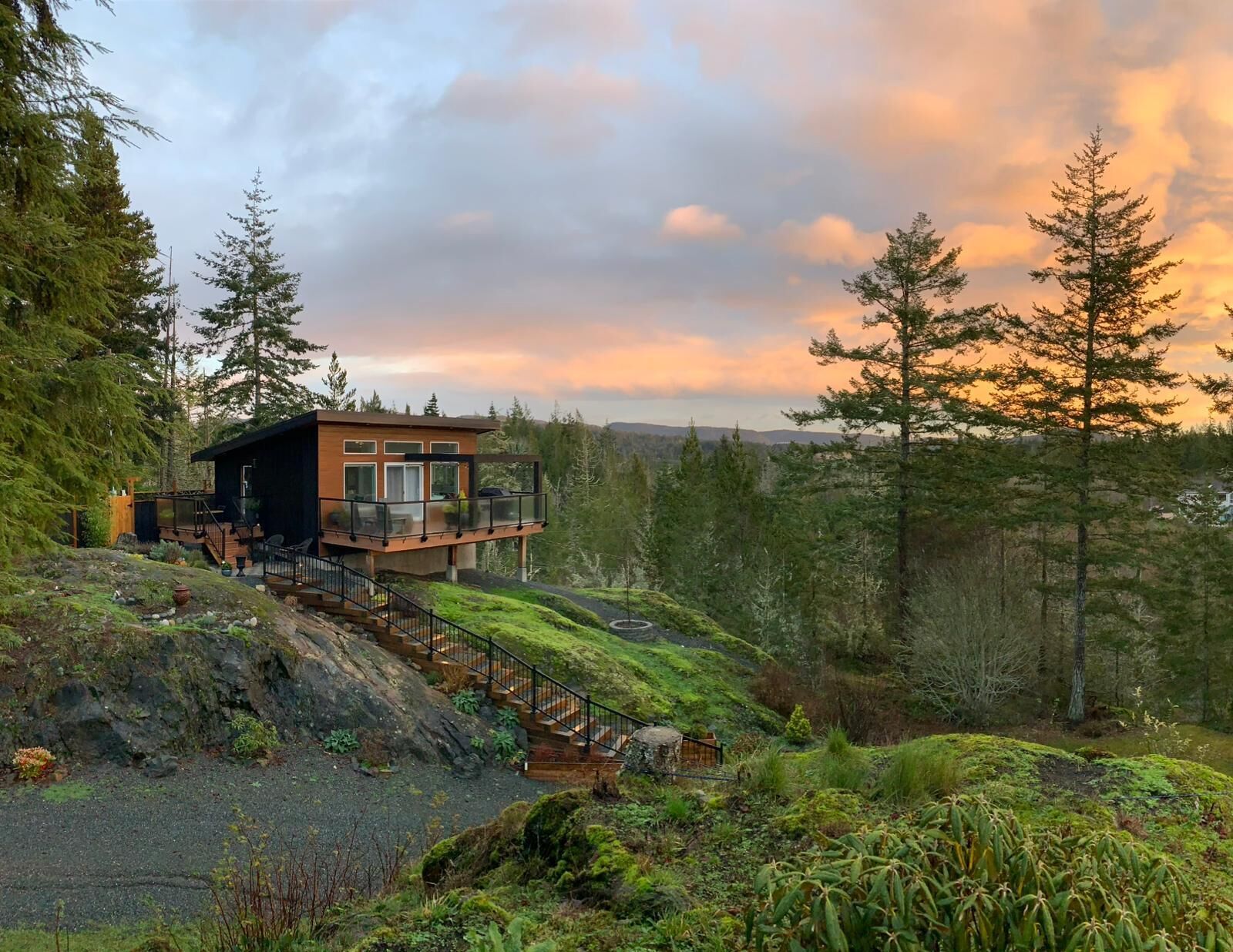 A modern cabin with a deck, surrounded by forest and mountains.