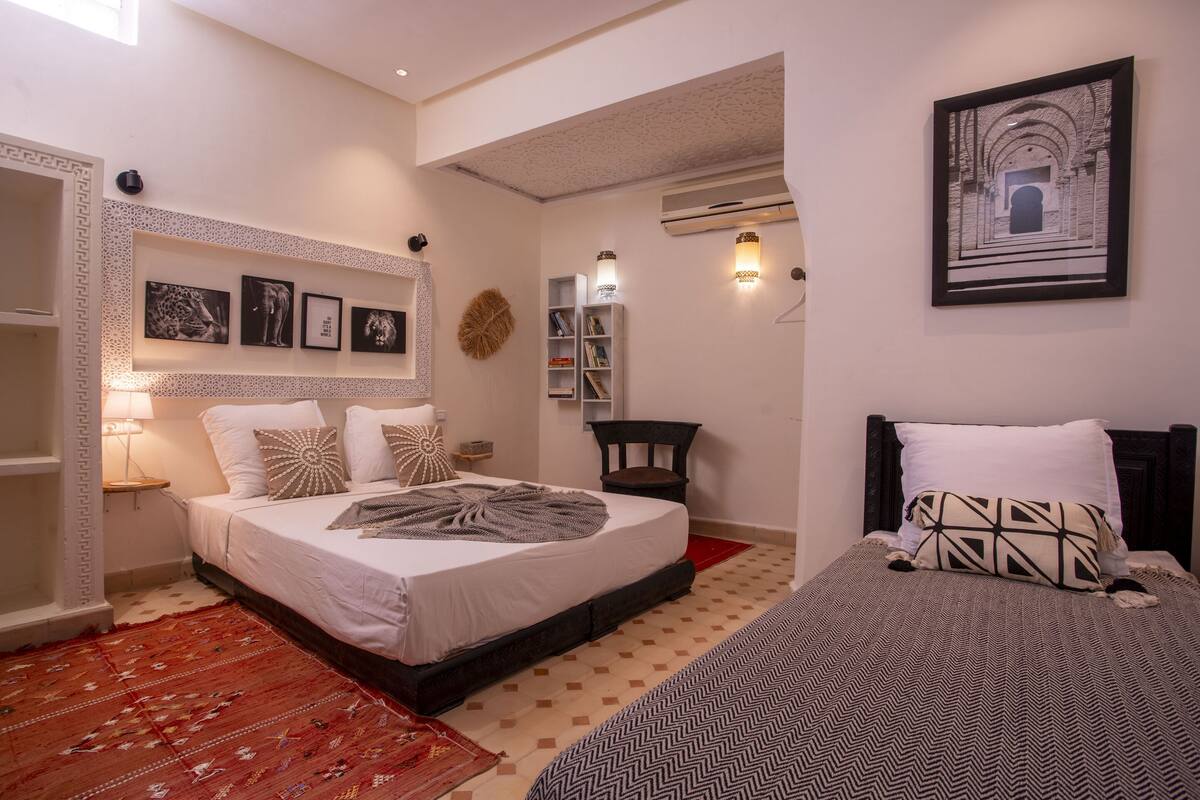A bedroom with a large bed, a side table, a lamp, a bookshelf, and a framed picture on the wall.