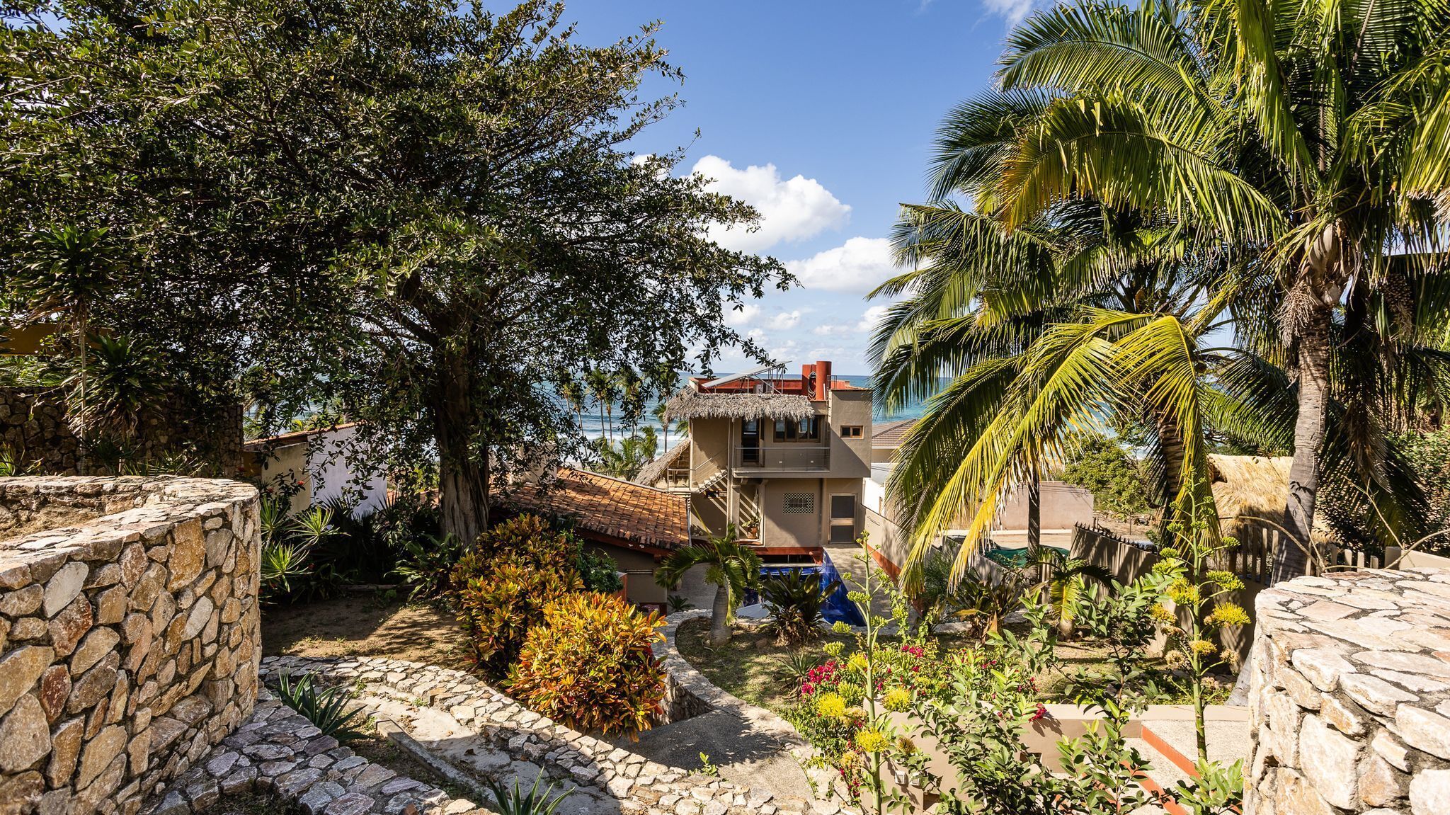 A stone-walled courtyard with a thatched-roof building, surrounded by lush greenery and palm trees.