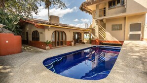 Una piscina con superficie de agua azul cristalina, rodeada por una zona de grava y un edificio con tejado de azulejos y un balcón.