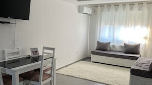Una sala de estar moderna con comedor, televisor de pantalla plana en la pared, sofá con cojines y un ventanal con cortinas translúcidas.