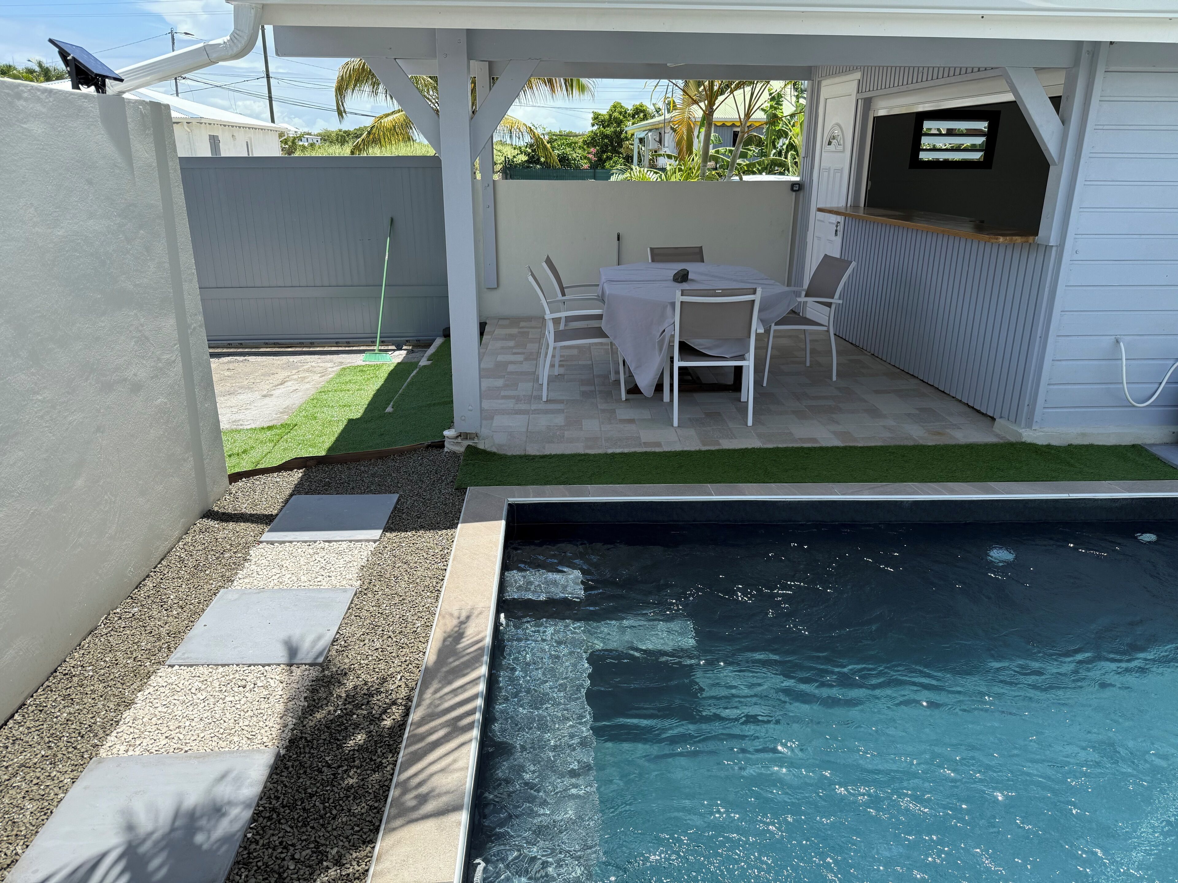 A backyard with a pool, a covered patio area with a table and chairs, and a small garden with artificial grass.