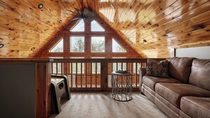 A cozy room with a sloped wooden ceiling, a brown leather sofa, a small round table, and a window with a view of trees.