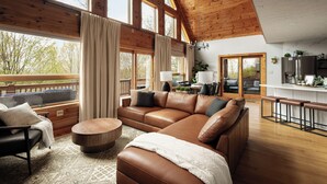 A spacious living room with a large brown leather sectional sofa, a round wooden coffee table, and a view of the outdoors through large windows.