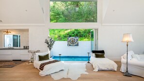 Ein modernes Schlafzimmer mit großem Fenster, einem Bett, einer Couch, einer Lampe und Blick auf einen Swimmingpool.