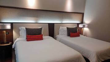 Habitación de hotel con dos camas, cada una con ropa blanca y una almohada roja decorativa. Hay dos lámparas de noche y una mesita de noche con una lámpara.