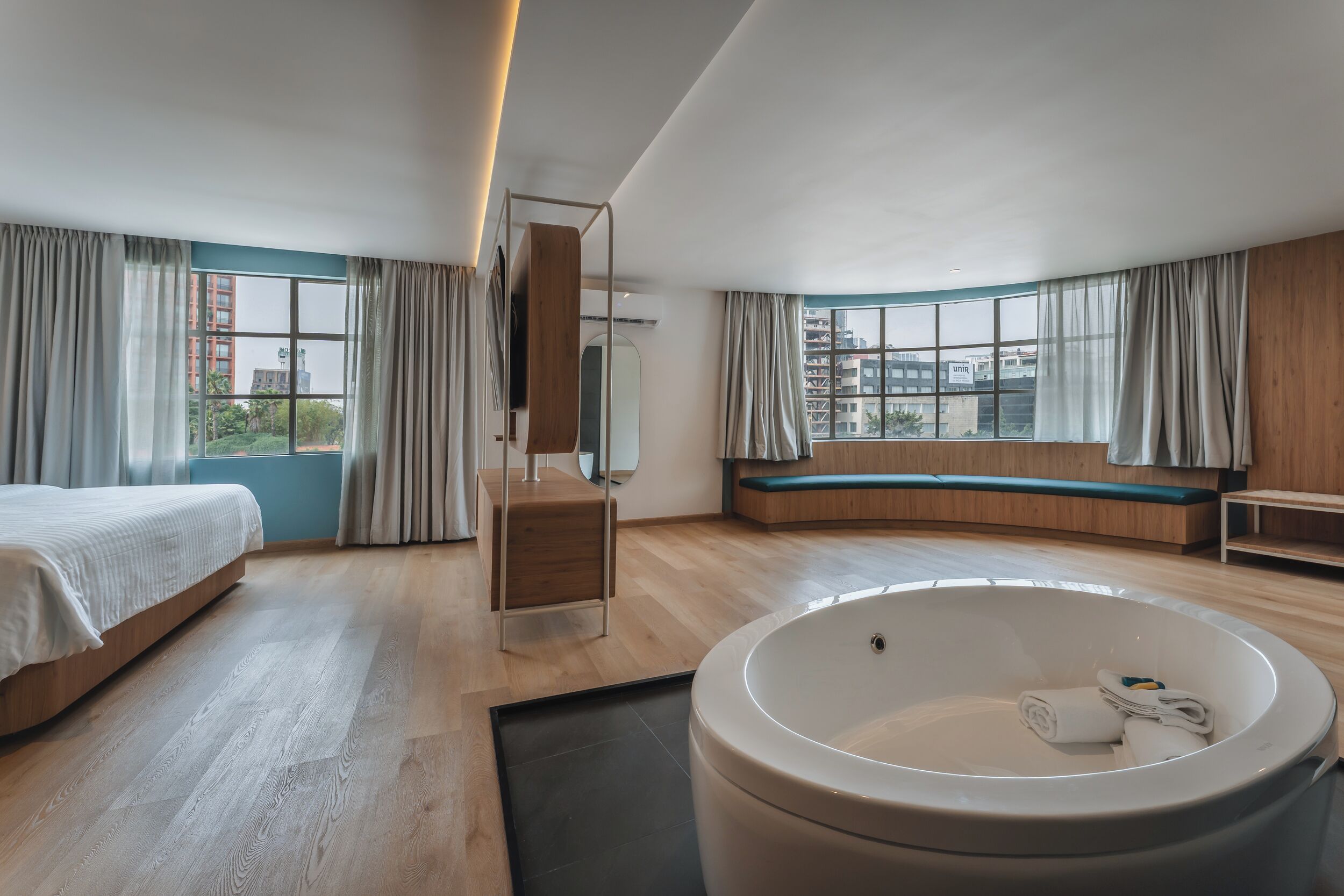 A modern hotel room with a large bathtub, a bed, a seating area, and a view of the city.