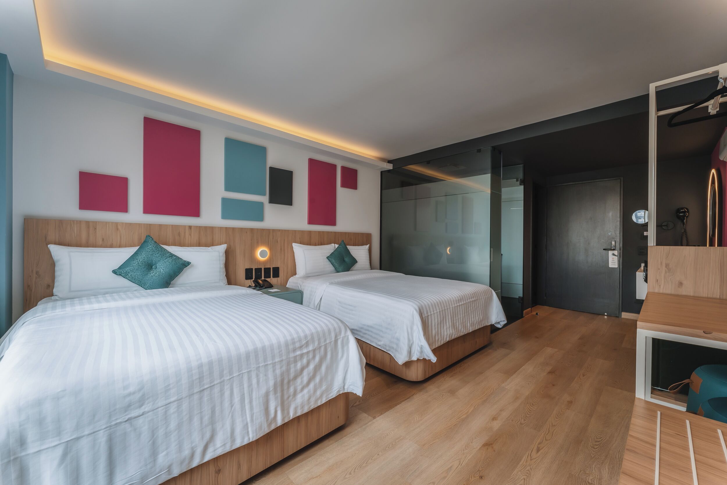 A hotel room with two beds, a wooden floor, a modern wall design, and a glass door leading to a bathroom.