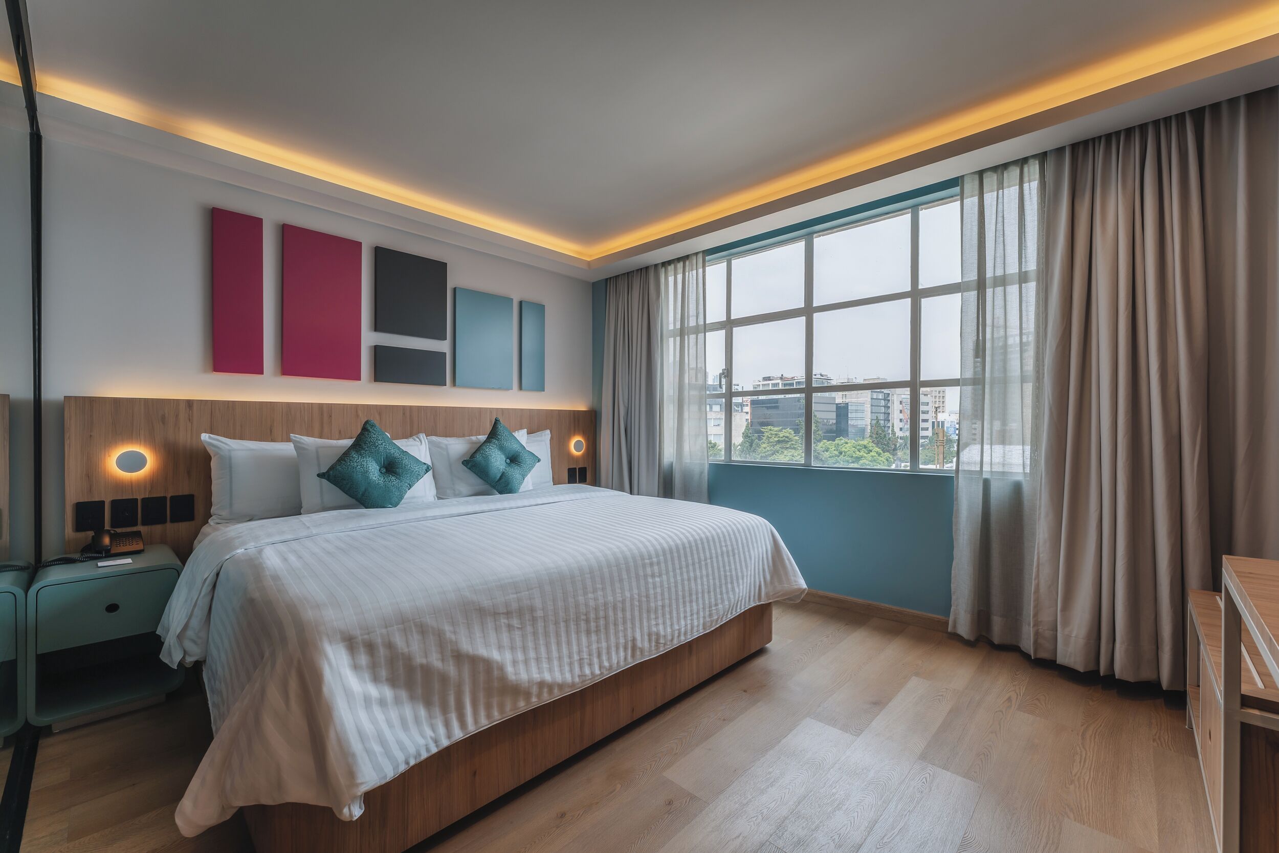 A modern hotel room with a large bed, a nightstand, a desk, and a view of the city through the window.