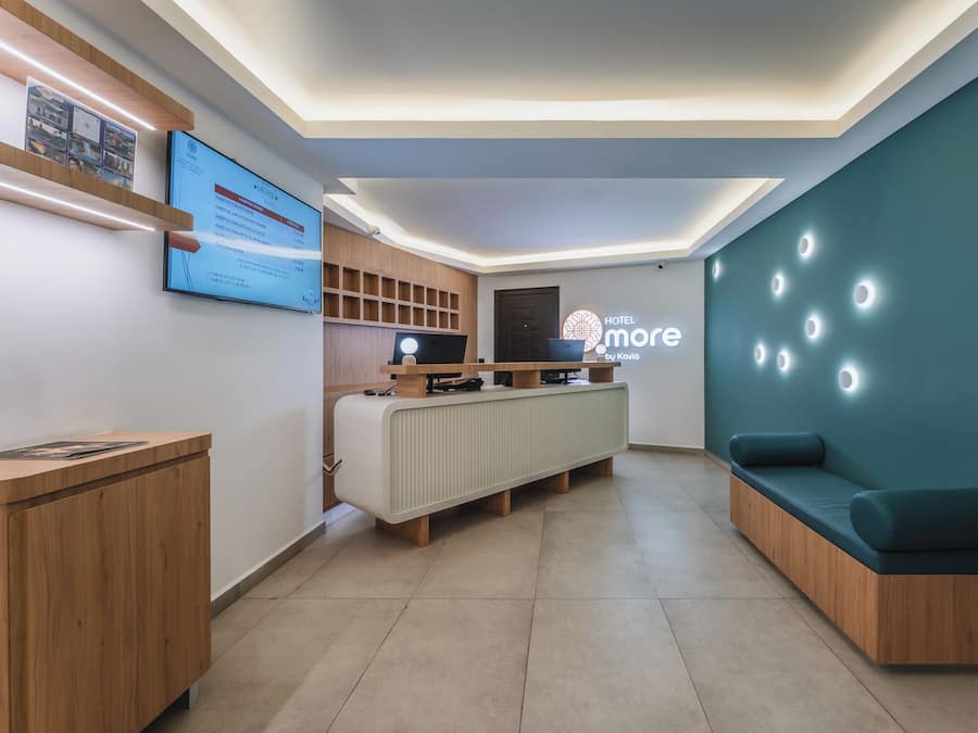 A modern hotel reception area with a digital display, wooden reception desk, and a seating area with a teal accent wall.