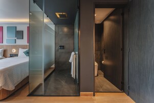 Une chambre d’hôtel moderne avec une cloison en verre, un lit, une table de chevet, une petite commode, une lampe fixée au mur et une petite étagère contenant des articles de toilette.