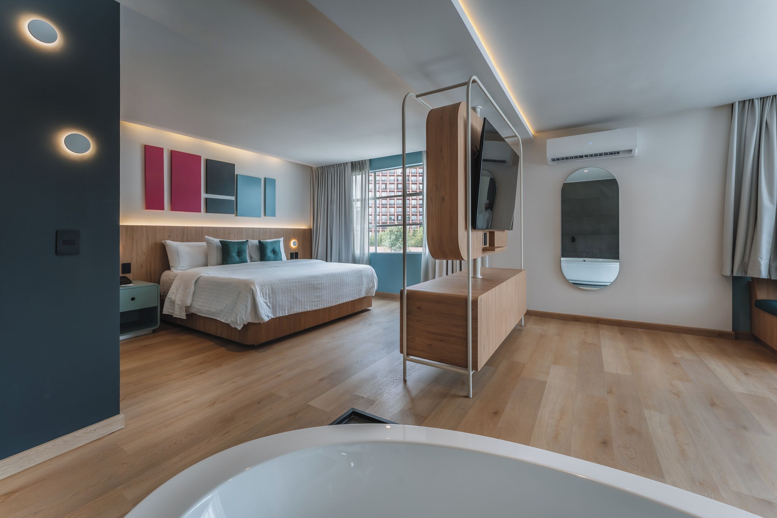 A modern bedroom with a large bed, a bedside table, a mirror, and a window with curtains.