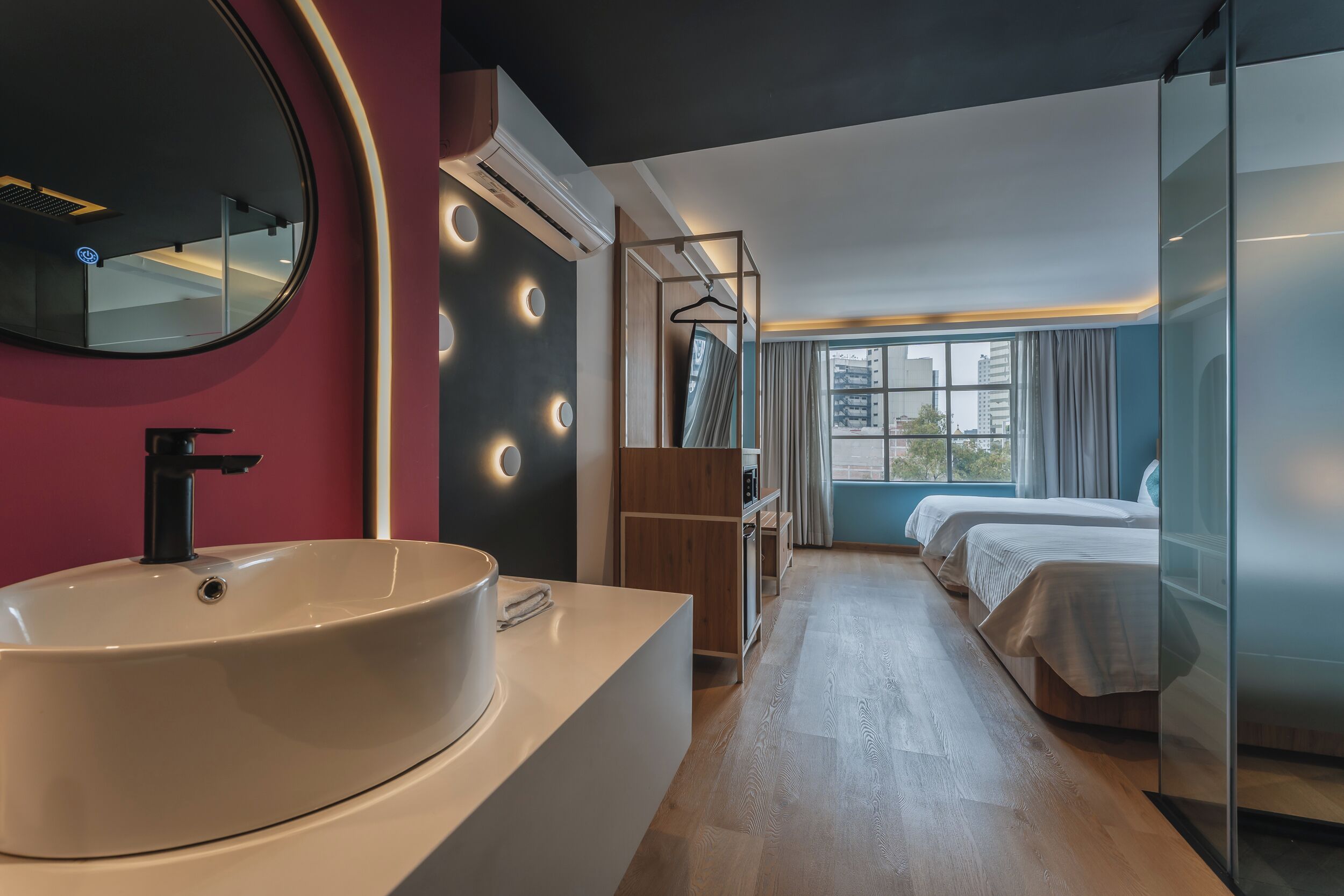 A modern hotel room with a large round sink, a mirror, a wardrobe, a bed, and a window with a view of buildings.