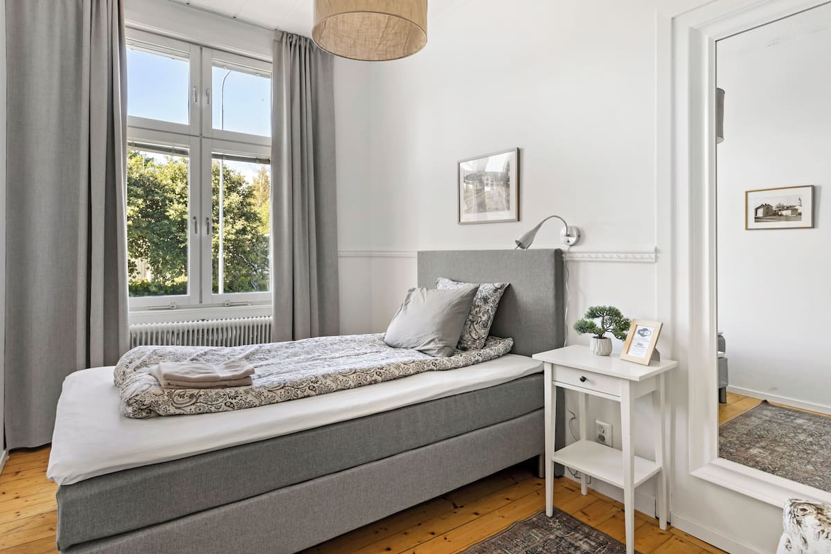 A bedroom with a bed, a bedside table, a radiator, a window with curtains, and framed pictures on the wall.