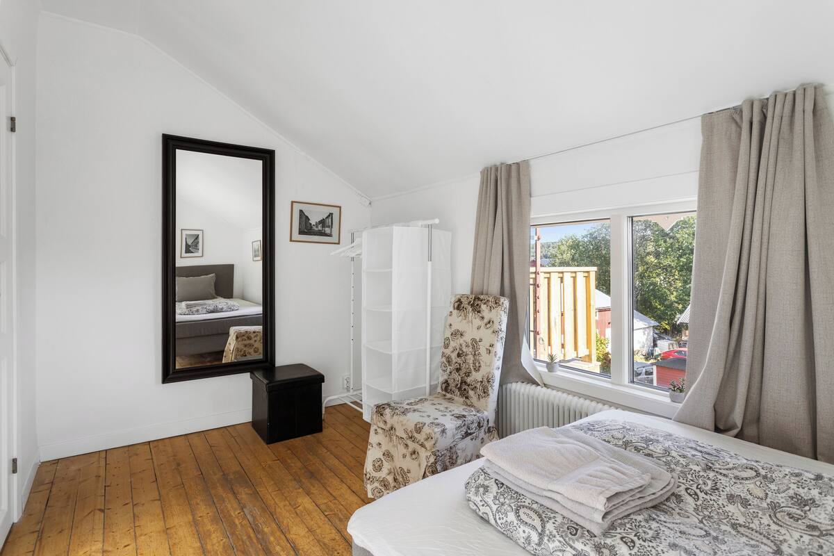 A bedroom with a large bed, a chair, a radiator, a window with curtains, and a mirror on the wall.