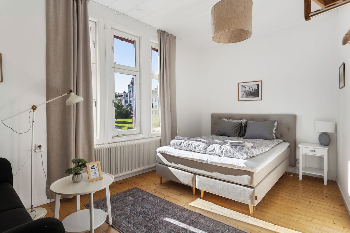 A bedroom with a bed, a bedside table, a lamp, a small table with a plant, and a view of the outside.