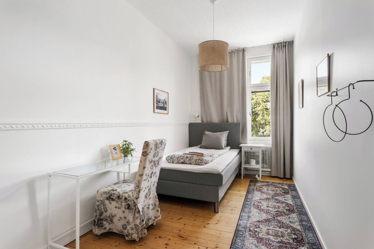 A modern bedroom with a grey bed, a white desk, a patterned chair, and a large window with curtains.