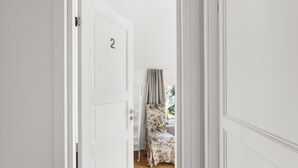 Un pasillo con una puerta blanca marcada con el número 2 que da a una habitación con una butaca de estampado floral y una ventana con cortinas.