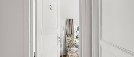 Ein Flur mit einer weißen Tür mit der Aufschrift '2', die zu einem Raum führt, in dem sich ein stufenartig gepolsterter Sessel und ein Fenster mit Vorhängen befinden.