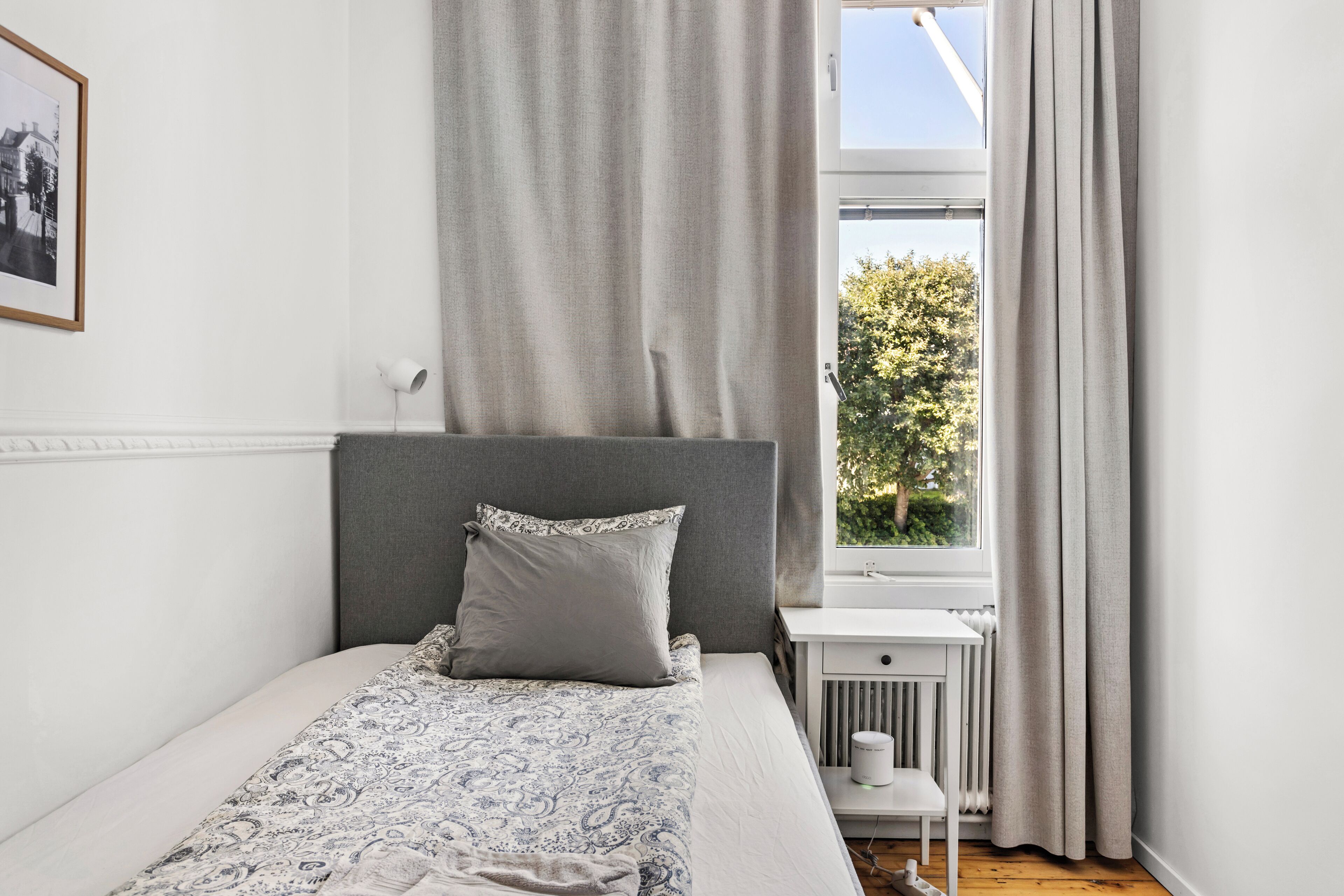 A neatly arranged bedroom with a bed, a bedside table, a radiator, and a window with curtains.