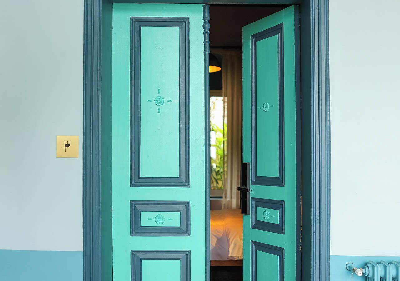 A teal door with a keyhole and a small lock, leading to a room with a bed and a window.