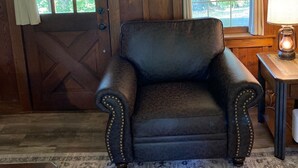 A rustic room with a leather armchair, wooden walls, a patterned rug, and a window with a view of trees. - Tarry Moore Cottage ... on Foxburg's historic fairways (Foxburg)