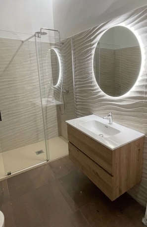 A modern bathroom with a glass shower enclosure, a wooden vanity with a sink, and a large mirror with a circular light. - Nerisa home  (Naples)