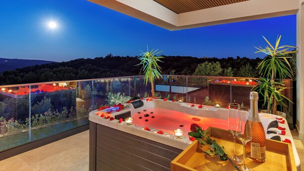 A hot tub with rose petals and candles, surrounded by plants, on a balcony with a view of the hills.
