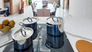 Una cocina con ollas de acero inoxidable sobre una encimera, un cuenco de frutas y una mesa de comedor al fondo.