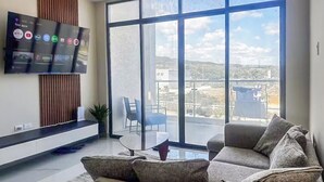 A modern living room with a large TV mounted on the wall, a sofa, a coffee table, and a view of the outside through large windows. - Paradisiac Beach Club 2BR | Pool, Beach & Balconies (st ann)