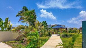 Un sendero de jardín que lleva a un pabellón con techo azul, rodeado de plantas tropicales y un cielo azul despejado.
