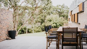 An outdoor patio with a long wooden table and chairs, surrounded by brick walls and greenery.