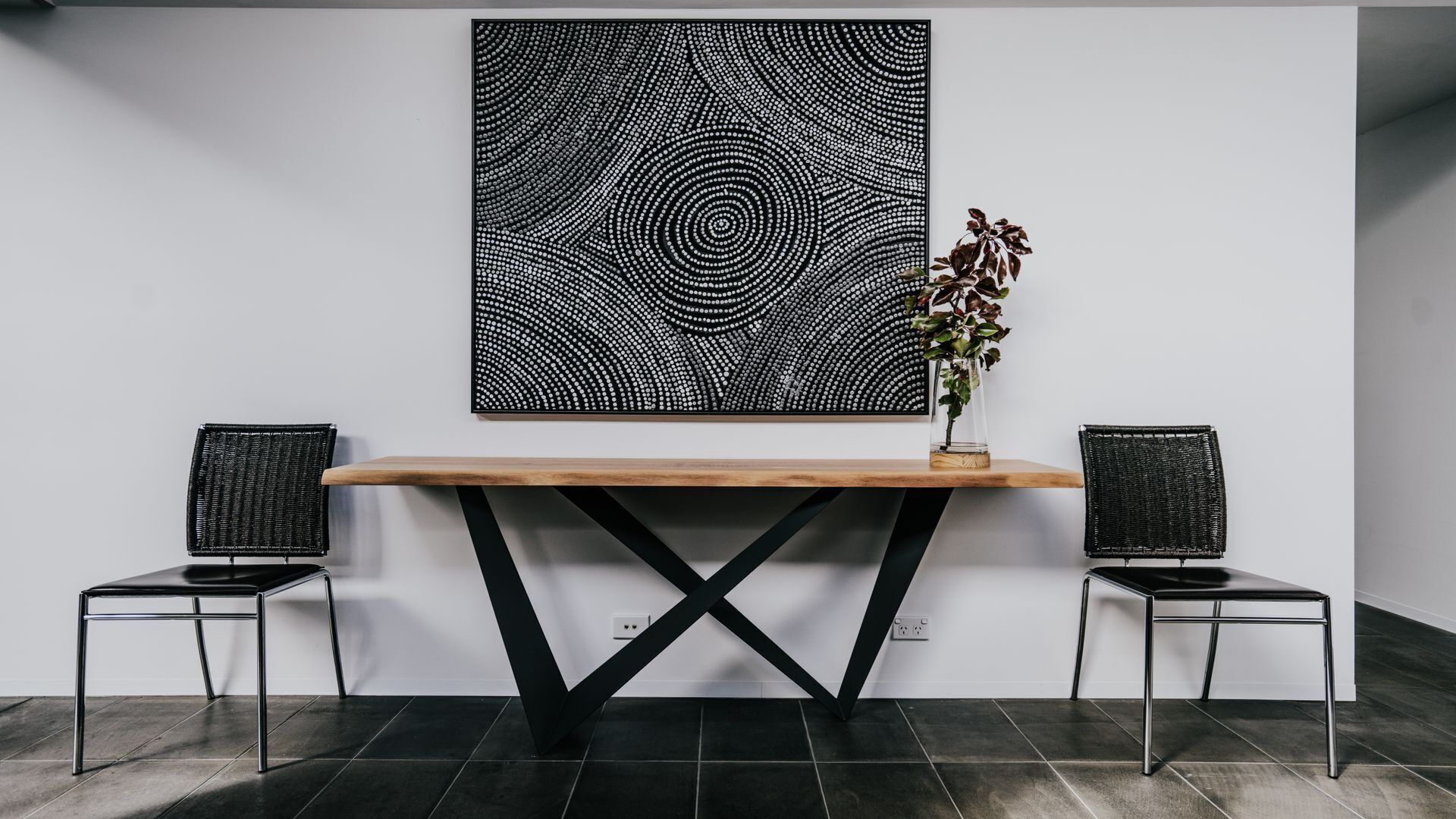 A reception desk with a wooden top and black metal legs, flanked by two black wicker chairs. A large black and white abstract artwork hangs above the desk.