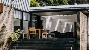 A modern outdoor seating area with a wooden table and chairs, a brick wall, and a glass door leading to an interior space. - Alkina Lodge Yani is the ultimate base for your Great Ocean Road adventure. (Wattle Hill)