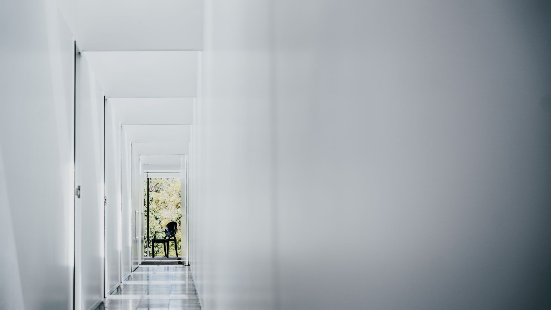 A narrow, white hallway with a door at the end leading to a room with a table and chair.
