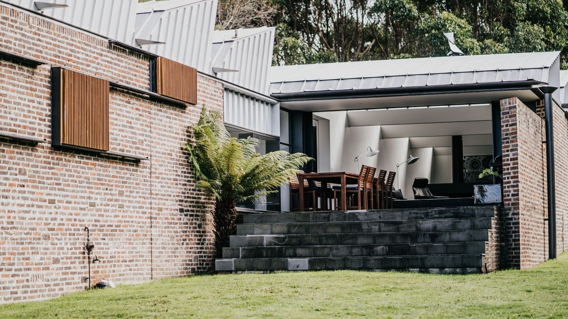 A modern house with a brick exterior, a covered outdoor seating area with a table and chairs, and a large fern plant.