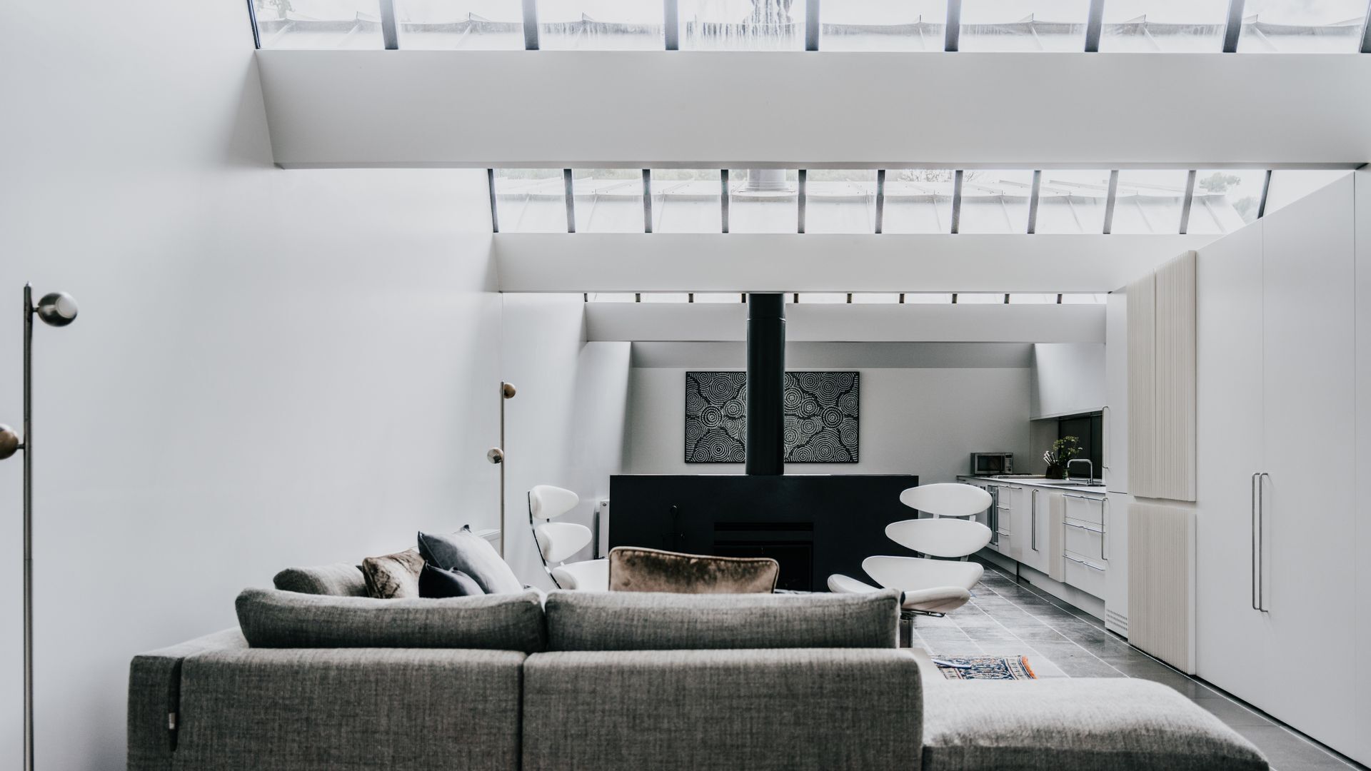 Un salon moderne avec un canapé gris, un meuble de rangement noir pour appareils électroniques et un coin cuisine doté de meubles blancs et d’un four à micro-ondes.
