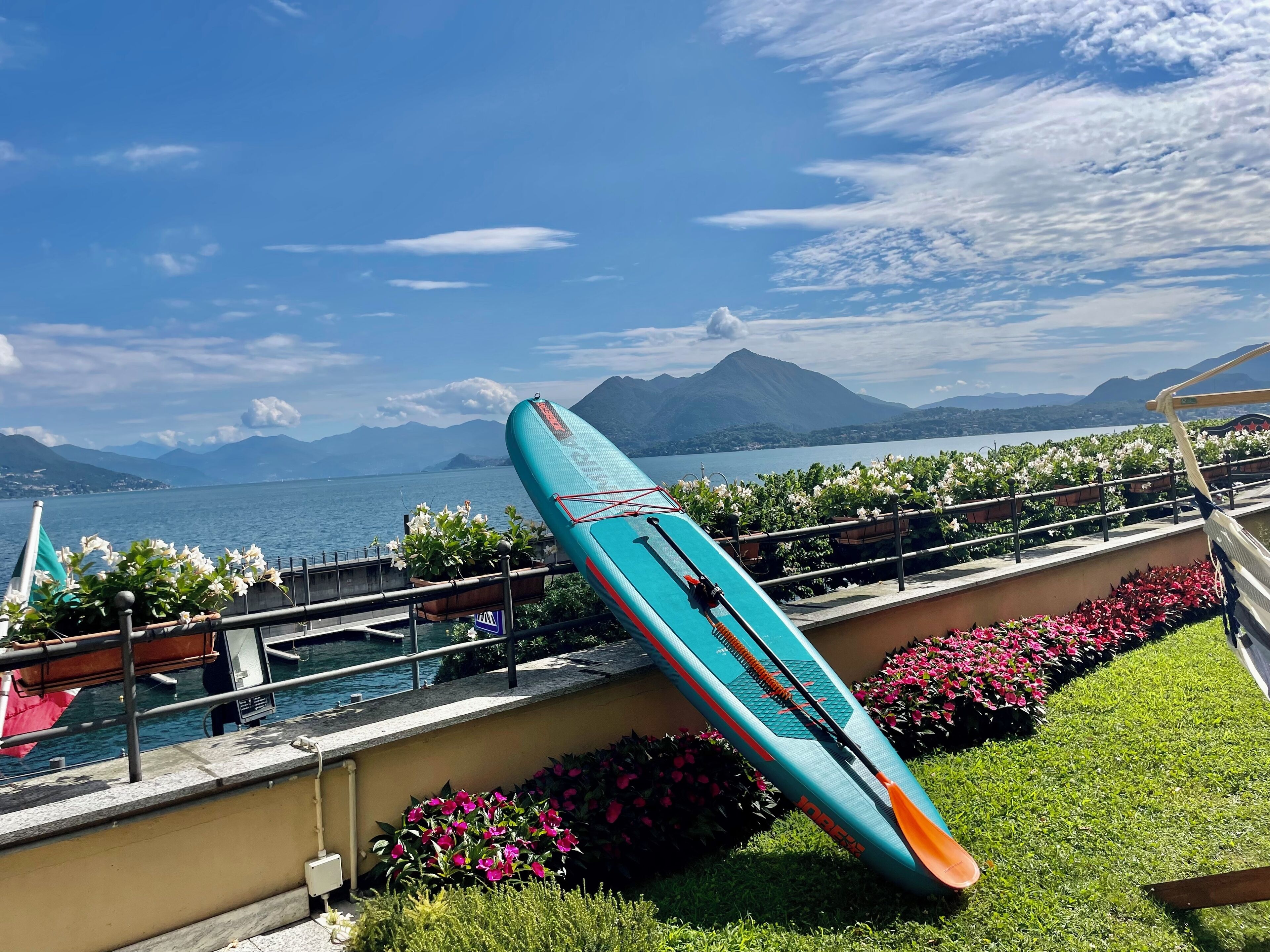 Un kayak apoyado en una barandilla junto a un paseo marítimo a la orilla de un lago, con montañas al fondo.