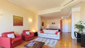Una sala de estar moderna con dos sofás rojos, un sofá blanco y una mesa de centro de madera. Hay un cuadro en la pared y una planta a la derecha.